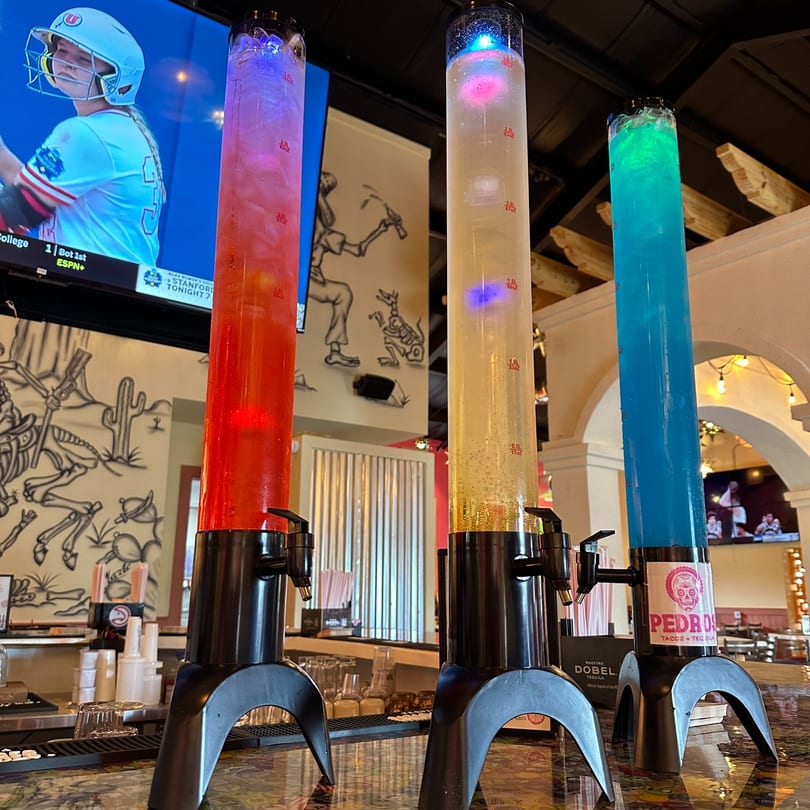 Three colorful Pedros Tacos & Tequila drink dispensers on a bar, with a TV showing sports in the background.