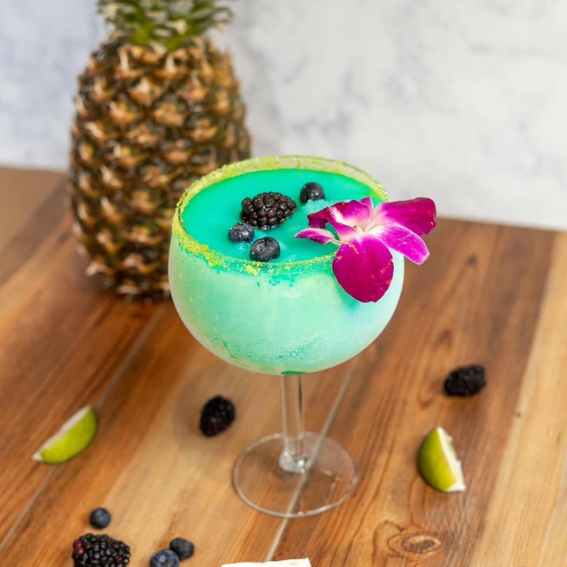 Teal margarita with berries and a pink orchid, next to a pineapple and lime wedges.