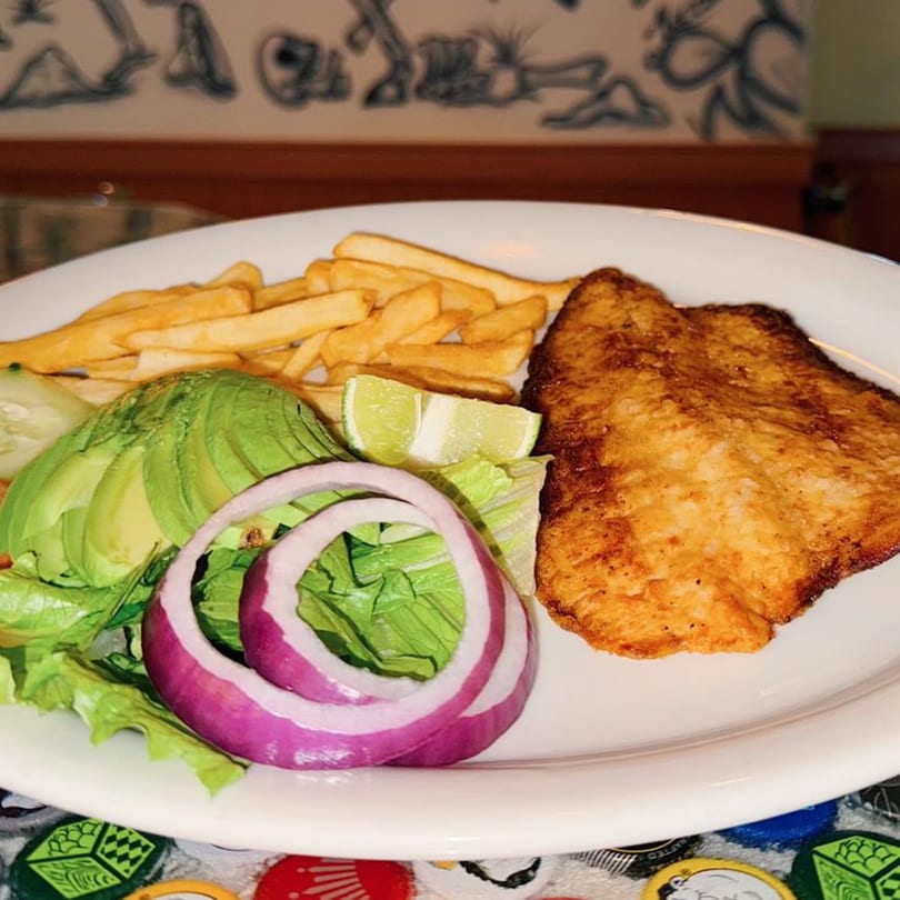 Fried fish fillet with french fries, avocado, red onion, lettuce, and lime on a white plate.