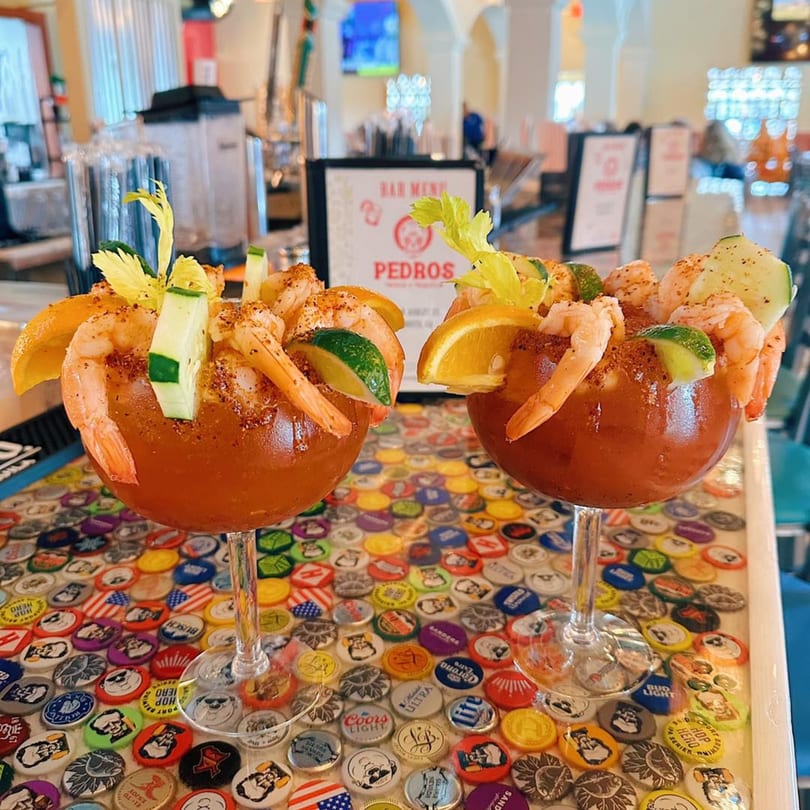 Two shrimp cocktails with celery, cucumber, and citrus on a colorful bottle cap bar top at Pedros.