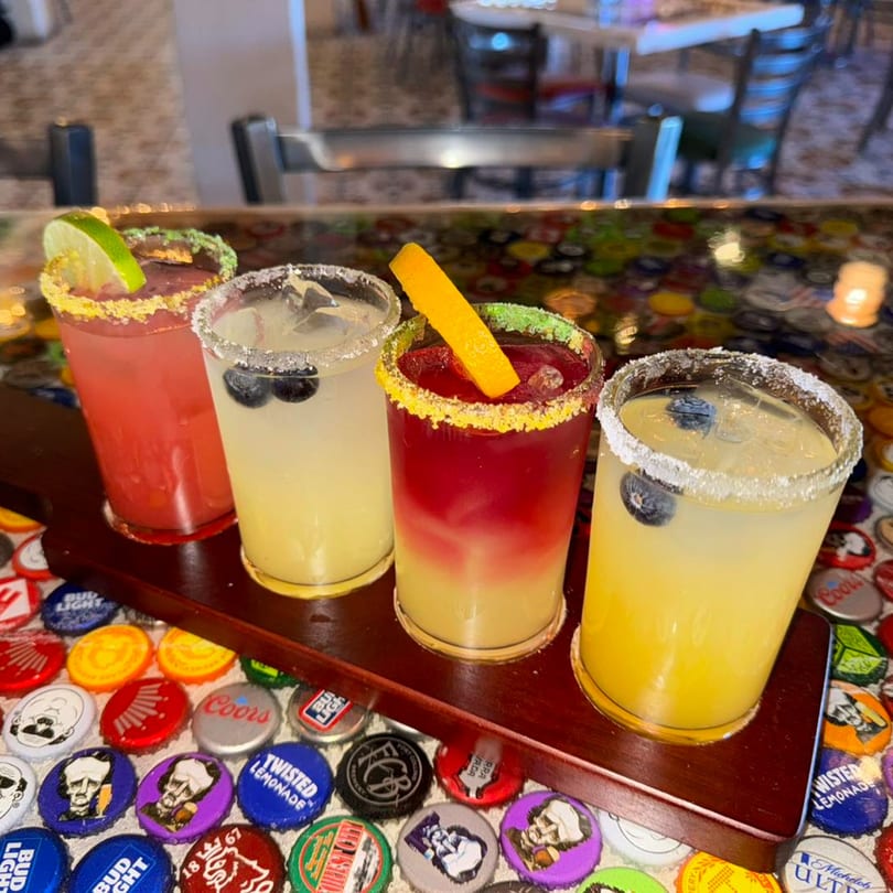 Four colorful margaritas with fruit garnishes on a wooden paddle, on a bottle cap covered table.