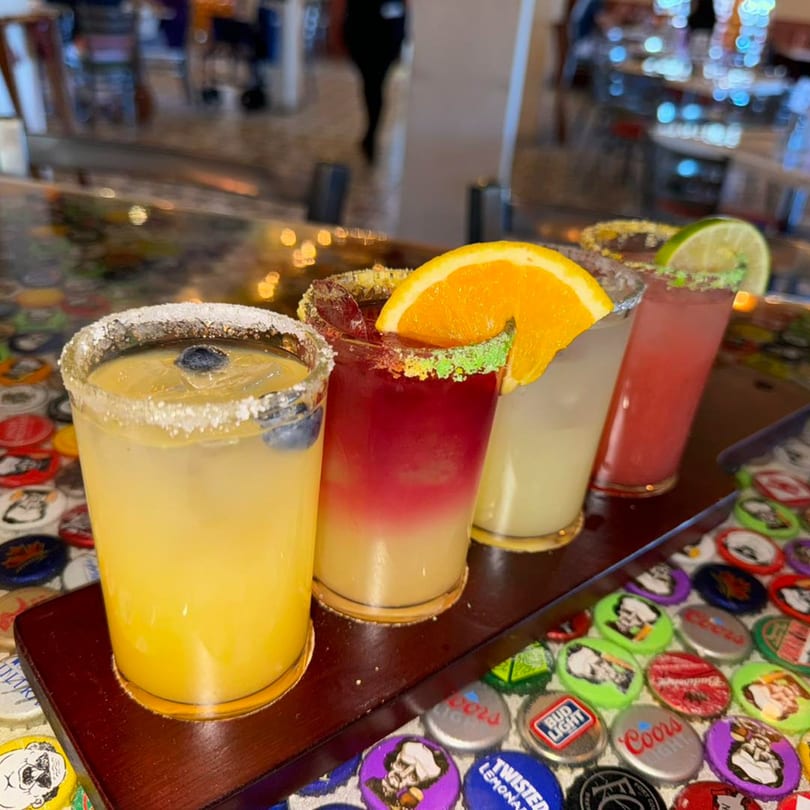 Four colorful Pedros Tacos & Tequila margaritas with fruit garnishes on a bottle cap table.