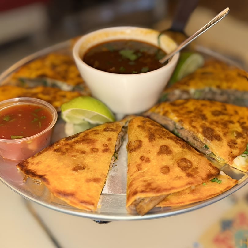 A platter of golden-brown quesadillas, with a side of red salsa, lime wedges, and a bowl of birria broth.