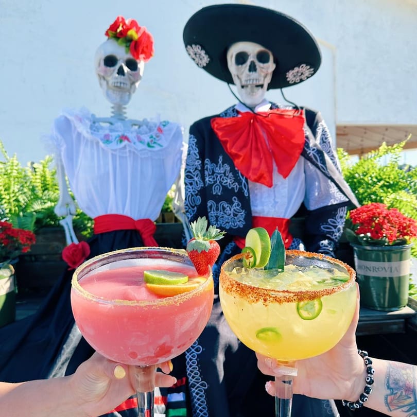 Two hands hold a strawberry and a spicy margarita in front of two festive skeleton decorations.
