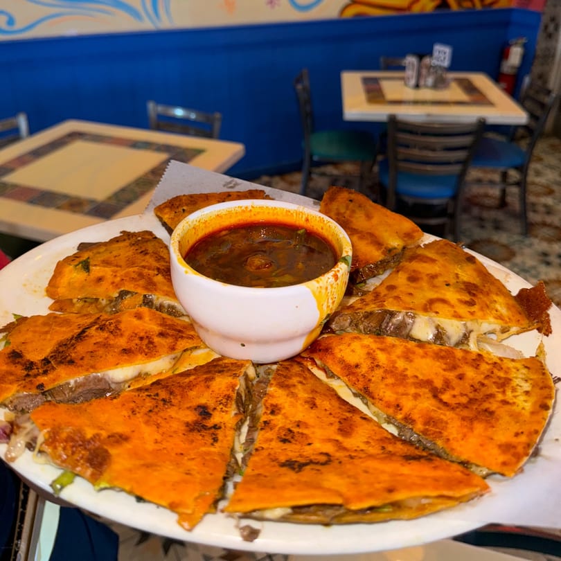 A plate of cheesy birria quesadillas with a side of consommé, served in a vibrant Mexican restaurant.