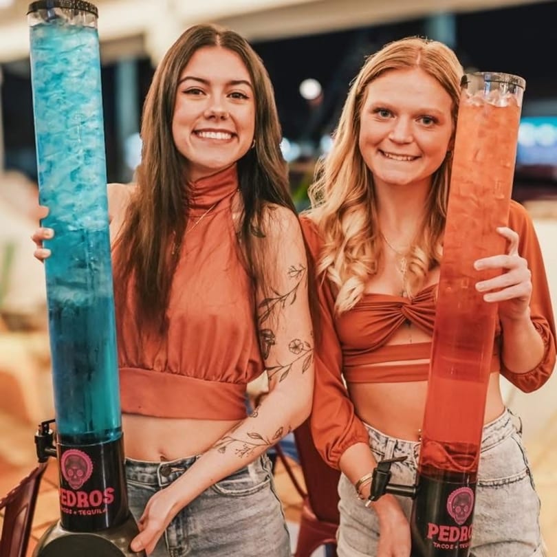 Two smiling women holding tall Pedros Tacos & Tequila drink dispensers, one blue and one red.